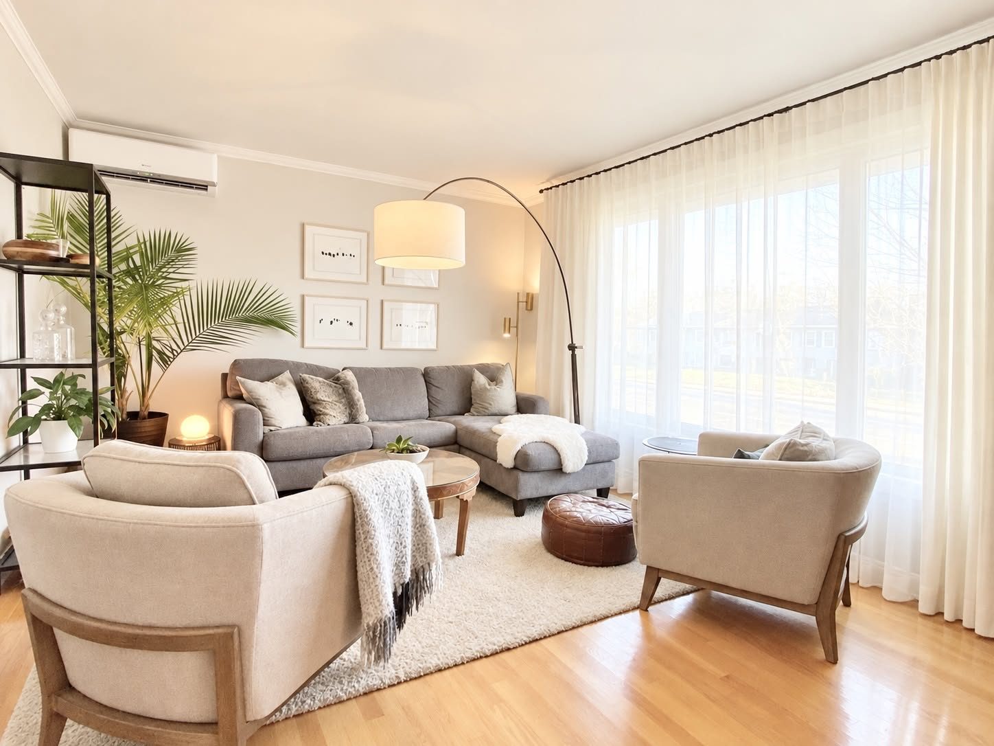 Salon lumineux : sofa sectionnel gris, fauteuils crème, rideaux voilage, lampe arquée et plantes vertes.